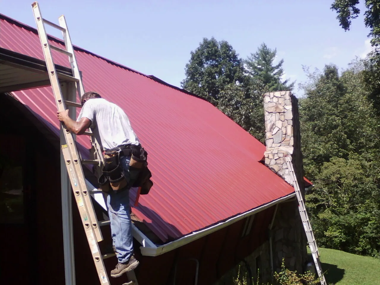 Roofer on a ladder beside a red metal roof and stone chimney — Soffit & Fascia Repair by Roofing Brothers in Molina
